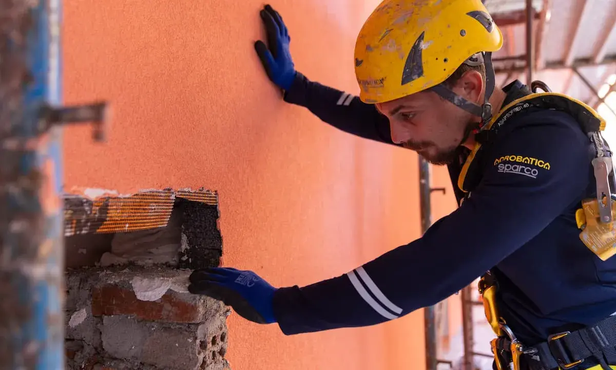 Un ouvrier en casque jaune effectue des travaux d'isolation extérieur, appliquant un enduit sur un mur orange.