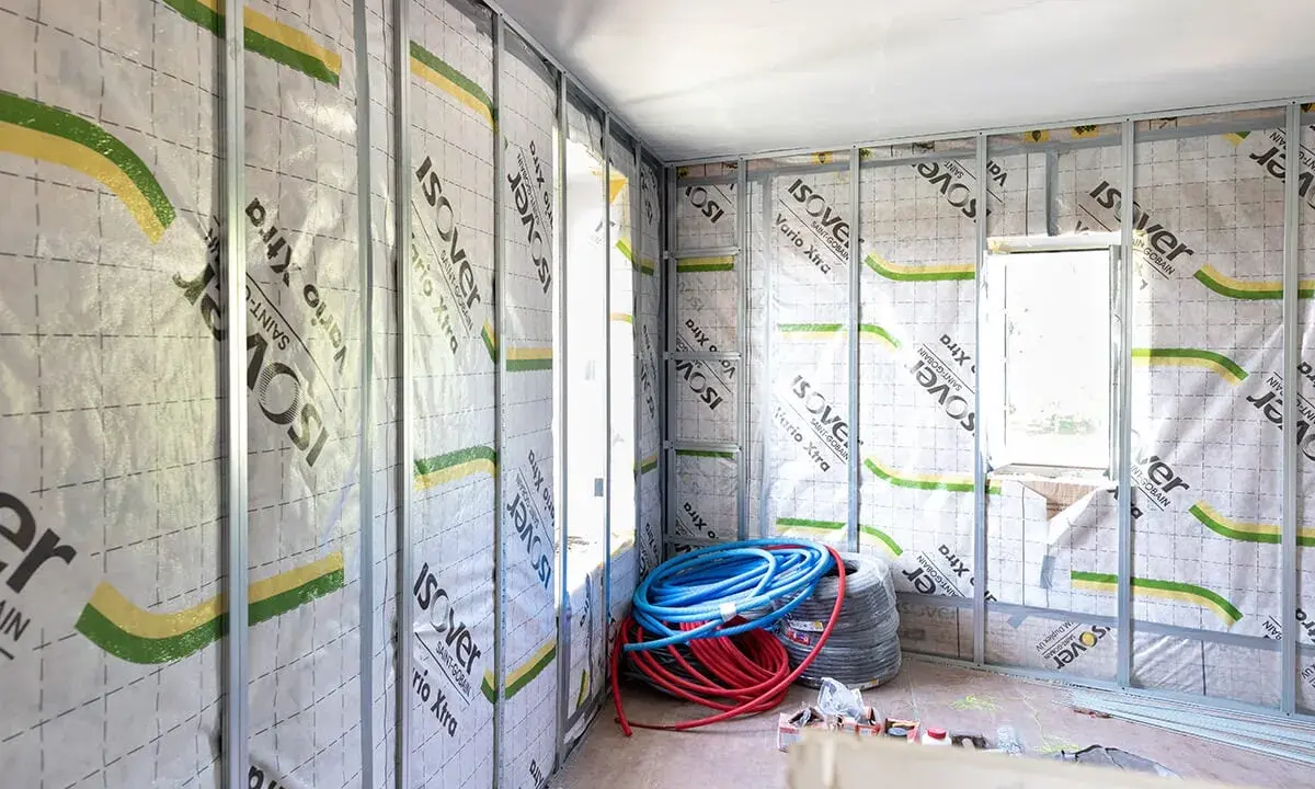 Intérieur en cours d'isolation, murs recouverts de pare-vapeur Isover Vario Xtra. Rouleaux de tuyaux bleus et rouges au sol. Un coup de pouce pour une isolation performante.