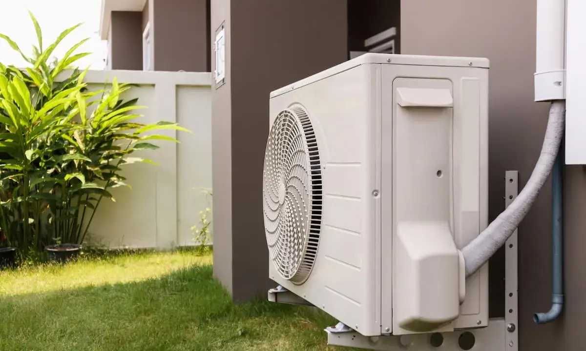 Unité extérieure de pompe à chaleur avec tuyau pour l'évacuation eau condensation. L'appareil est fixé au mur d'une maison, près d'une pelouse et de végétation.