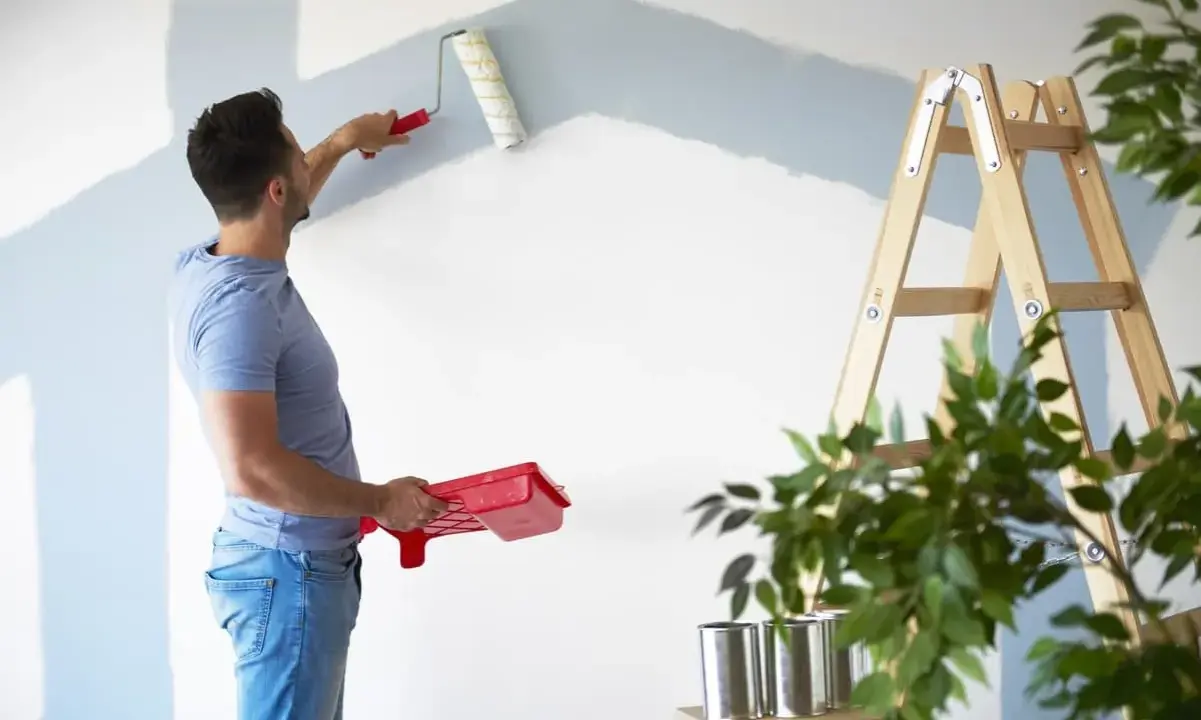 Homme peignant un mur en bleu clair, prêt à refuser assurance éco-ptz pour son projet de rénovation. Échelle et plantes à proximité.