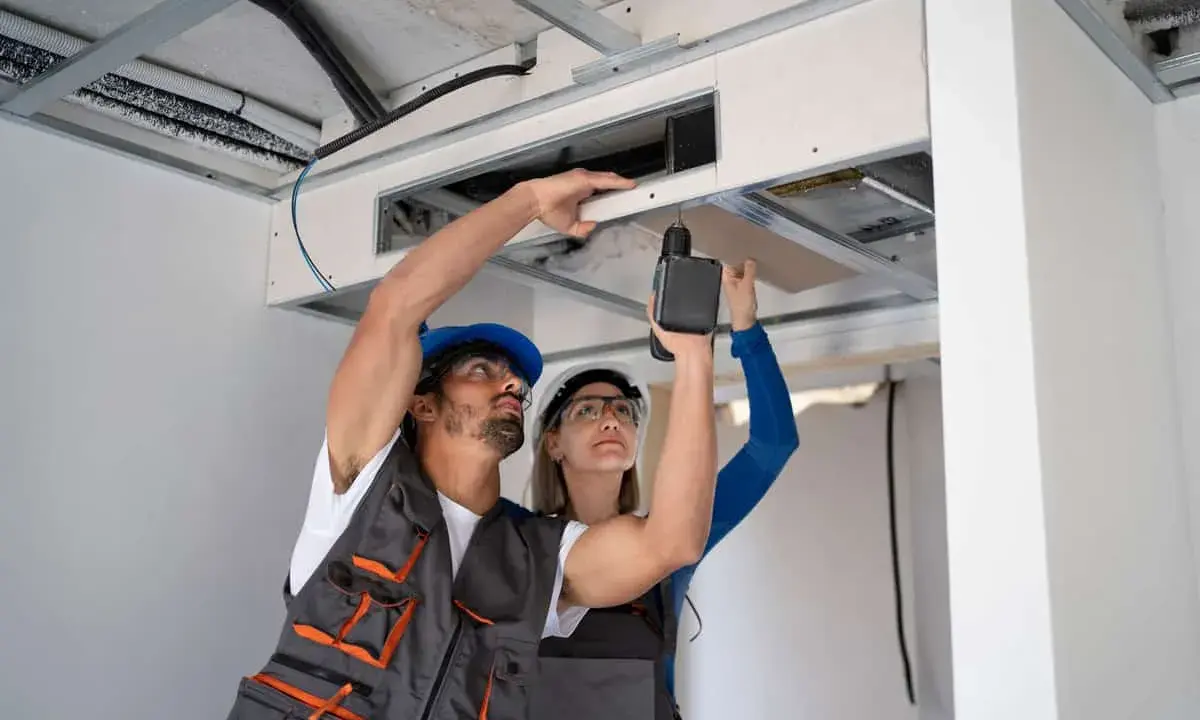 Deux ouvriers travaillent sur l'installation vmc simple flux, l'un perce le plafond, l'autre tient une plaque.