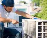 Un technicien effectue l'entretien d'une pompe à chaleur sur un toit, utilisant un tournevis pour ajuster l'unité extérieure.