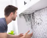 Un homme découvre de la moisissure sous un meuble de cuisine. Une VMC salle de bain norme est essentielle pour éviter ce problème.
