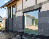 Maison en construction avec isolation par l'extérieur en panneaux gris. Les fenêtres reflètent le ciel bleu et un autre bâtiment.