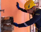 Un ouvrier en casque jaune effectue des travaux d'isolation extérieur, appliquant un enduit sur un mur orange.