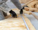 Pose d'isolant pour optimiser la résistance thermique du bois. Un ouvrier mesure et découpe la laine de verre.