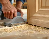Un artisan utilise une ponceuse pour le détalonnage d'une porte en bois, créant des copeaux fins sur l'établi.