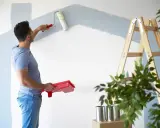 Homme peignant un mur en bleu clair, prêt à refuser assurance éco-ptz pour son projet de rénovation. Échelle et plantes à proximité.