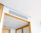 Grille de ventilation discrète pour un système de chauffage gainable, intégrée au plafond blanc et encadrée de bois.