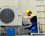 Technicien en bleu, casque jaune, installant une unité extérieure de pompe à chaleur. La garantie pompe à chaleur est assurée par ce professionnel.