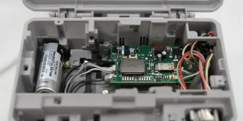 Intérieur d'un boîtier gris ouvert, révélant un circuit imprimé VMC, une batterie et des fils. Le VMC qui bip pourrait indiquer un problème.