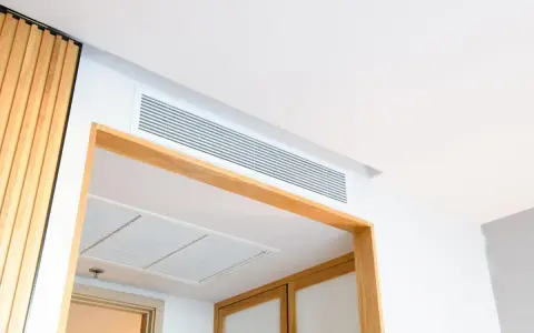 Grille de ventilation discrète pour un système de chauffage gainable, intégrée au plafond blanc et encadrée de bois.