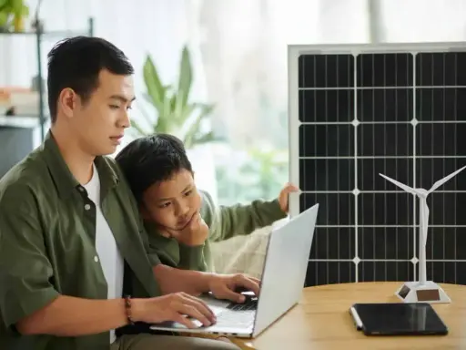 Père et fils étudient l'énergie solaire et éolienne sur un ordinateur portable, un panneau solaire et une éolienne à proximité. Un projet d'installation cacsi sans consuel.