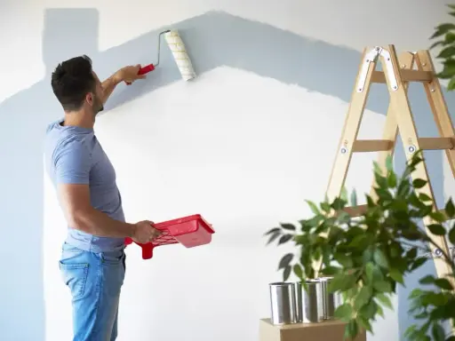 Homme peignant un mur en bleu clair, prêt à refuser assurance éco-ptz pour son projet de rénovation. Échelle et plantes à proximité.