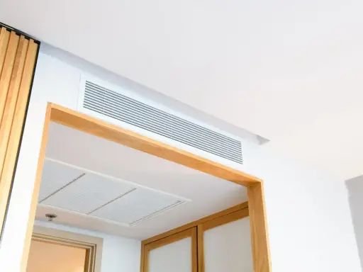 Grille de ventilation discrète pour un système de chauffage gainable, intégrée au plafond blanc et encadrée de bois.