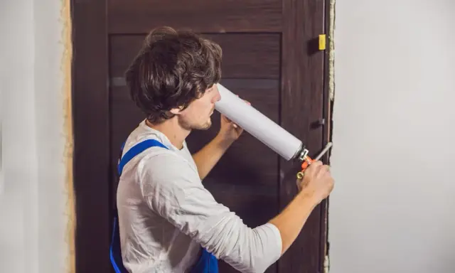 Un homme en salopette bleue utilise une mousse expansive pour comment isoler une porte.