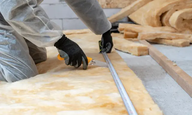 Pose d'isolant pour optimiser la résistance thermique du bois. Un ouvrier mesure et découpe la laine de verre.