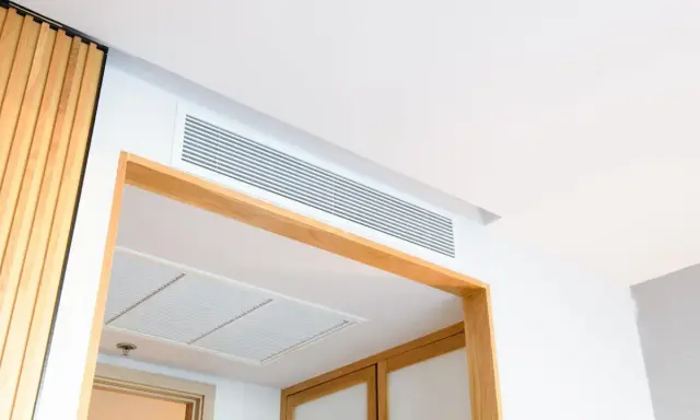 Grille de ventilation discrète pour un système de chauffage gainable, intégrée au plafond blanc et encadrée de bois.