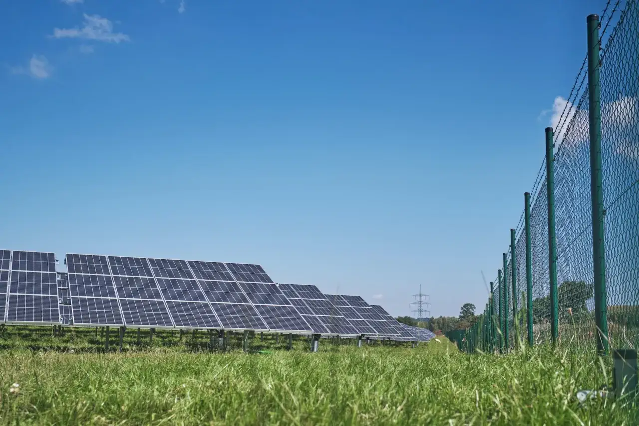 Lignes de panneaux solaires photovolta&iuml;ques sous un ciel bleu clair, bord&eacute;es par une cl&ocirc;ture grillag&eacute;e.