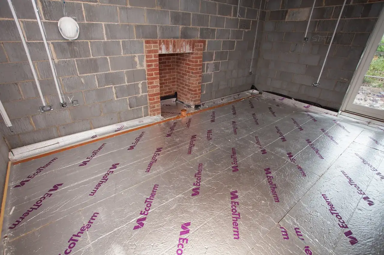 Chantier : pose de l'isolation sous carrelage dans une pièce aux murs en parpaings, avec une cheminée en briques.
