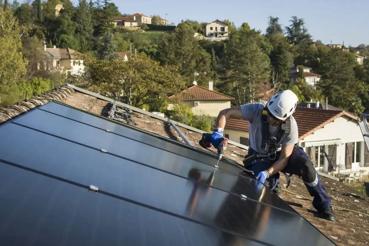 Un homme en casque et harnais installe des panneaux solaires sur un toit. Il utilise une perceuse pour fixer les &eacute;l&eacute;ments, une &eacute;tape cl&eacute; pour apprendre comment installer des panneaux solaires soi m&ecirc;me.