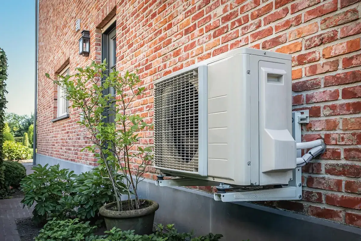 Unité extérieure d'une pompe à chaleur air air non réversible, fixée sur un mur de briques rouges, avec une plante verte à proximité.