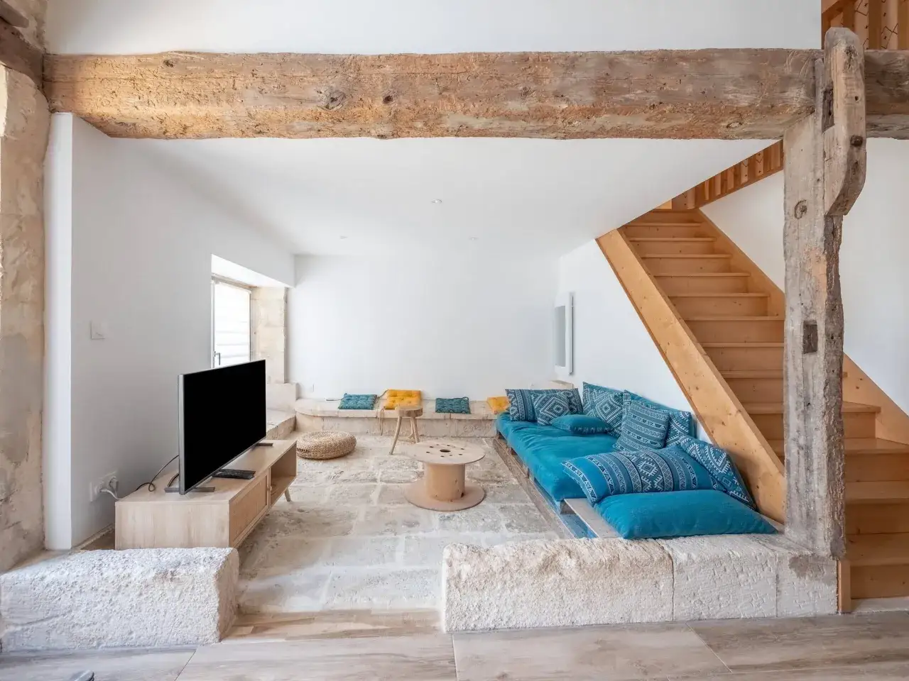 Salon r&eacute;nov&eacute; avec poutres apparentes, un canap&eacute; bleu, et un escalier en bois. Une belle aide r&eacute;novation maison ancienne.