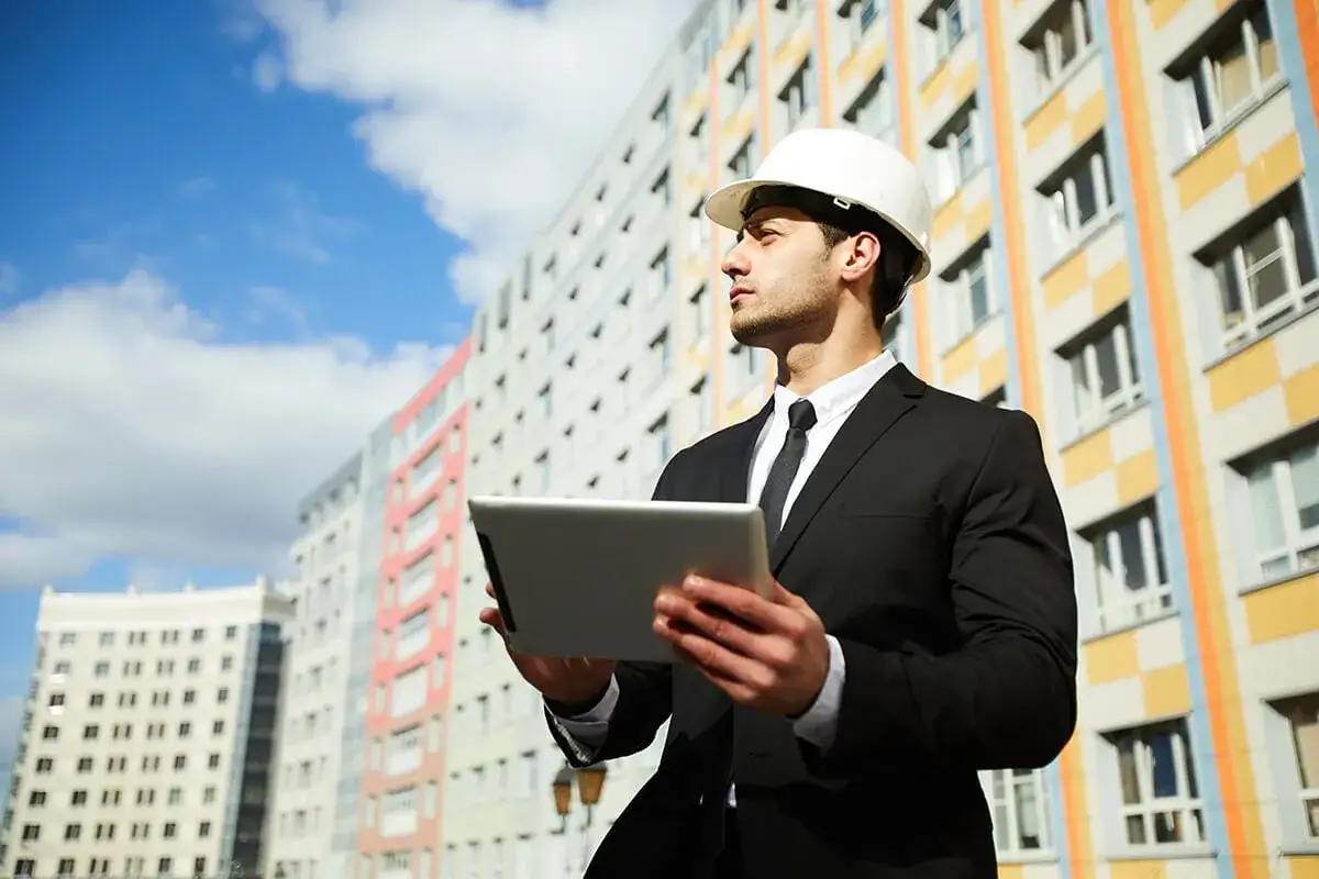Ing&eacute;nieur en costume et casque blanc, tablette en main, devant des immeubles color&eacute;s. Il pr&eacute;pare l'audit &eacute;nerg&eacute;tique obligatoire copropri&eacute;t&eacute;.