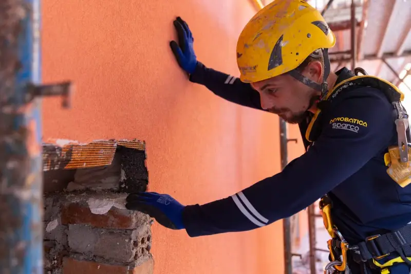 Un ouvrier en casque jaune effectue des travaux d'isolation extérieur, appliquant un enduit sur un mur orange.