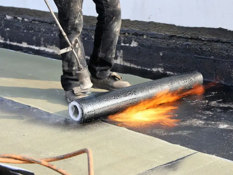 Un couvreur utilise un chalumeau pour souder un rouleau de membrane bitumineuse, créant ainsi une toiture chaude et étanche.